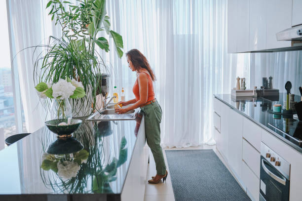 A woman is washing dishes in a modern, sunlit Dubai apartment kitchen with tall plants and sleek decor.