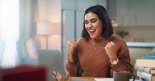 A woman in a brown sweater smiles while clenching her fists in excitement in front of a laptop.