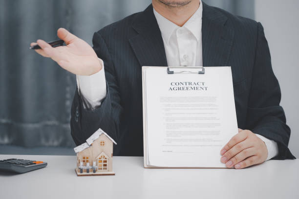 A man in a suit holds a contract agreement next to a small house model.