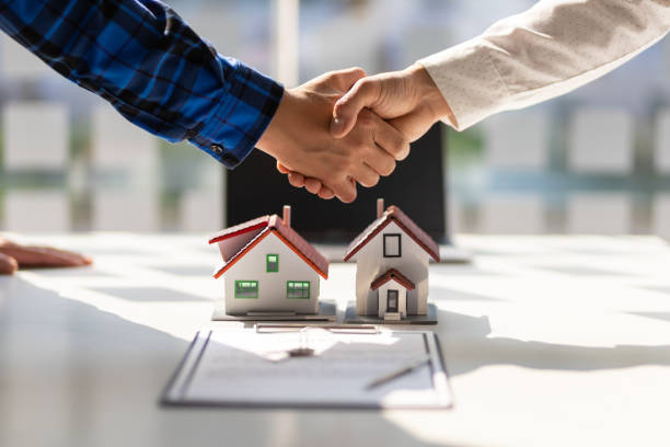 Two people shaking hands over a desk with small house models and a contract.