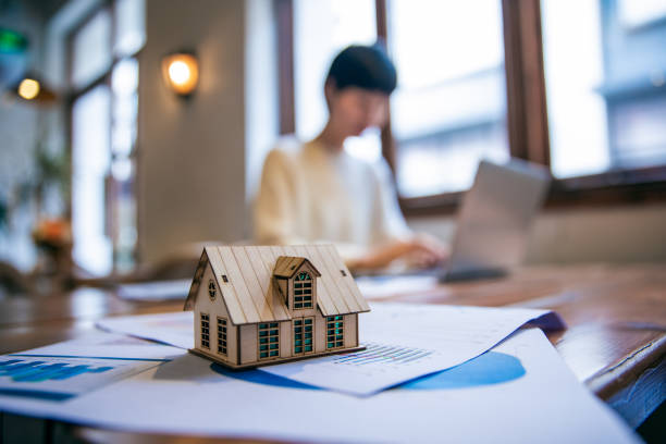 A small wooden house model sits on a table with documents, while a person works on a laptop in the background.