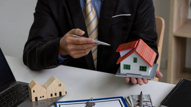 A person in a suit holding a small model house while pointing at it with a pen.